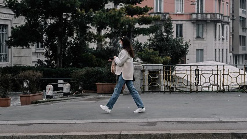 A woman in a face mask walks on a city sidewalk wearing denim jeans, embodying modern urban life.